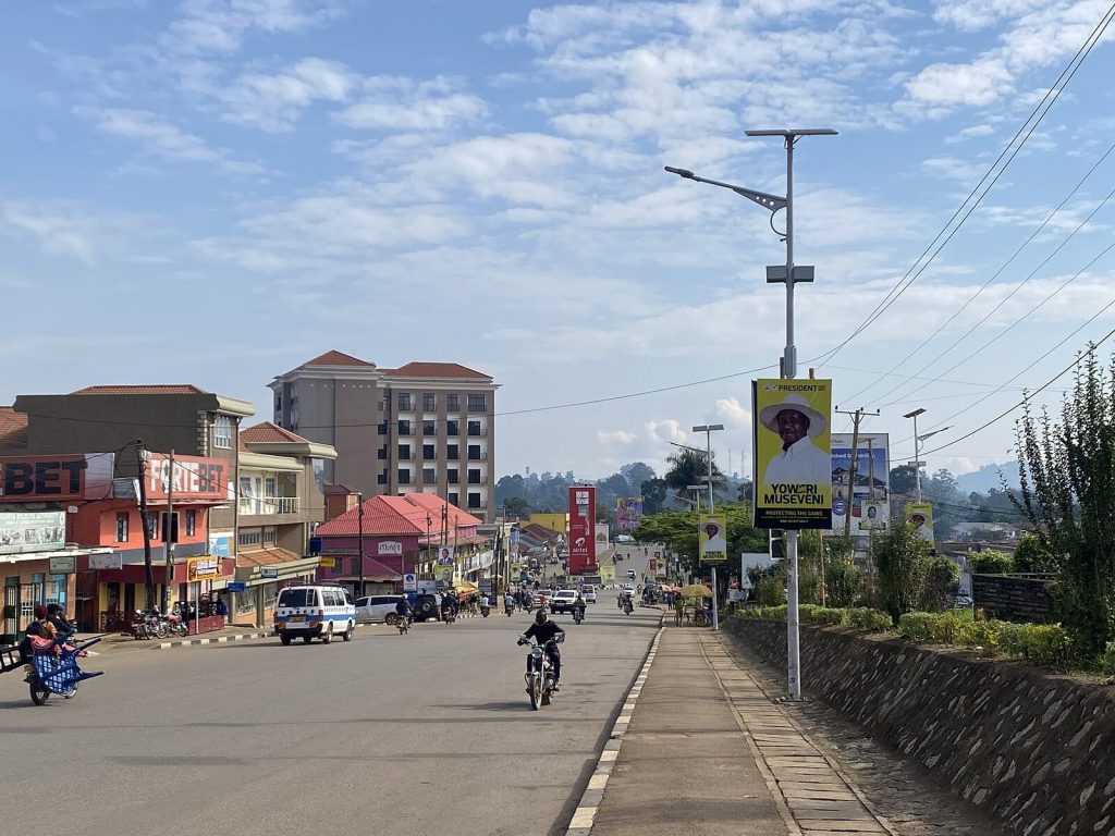 A main street in Fort Portal city looking downhill — shopfronts, boda-bodas, a matatu, solar