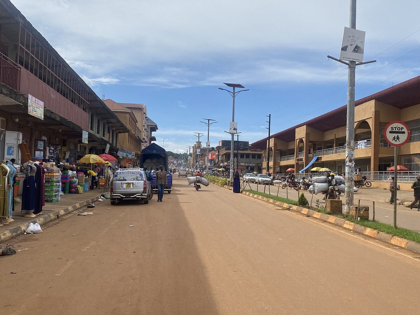 A broad central Masaka street with a planted median — long arcaded shop terraces on the left