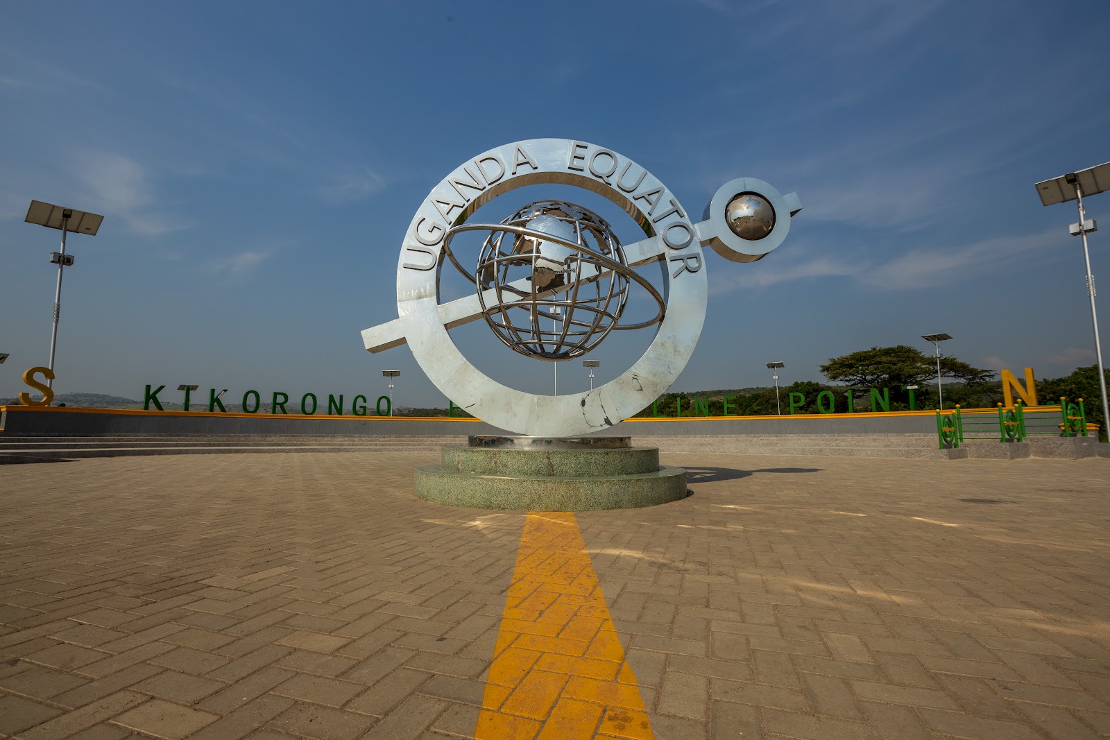 The steel Uganda Equator monument on its paved plaza at the Kikorongo equator line point, a yellow line marking the equator across the paving