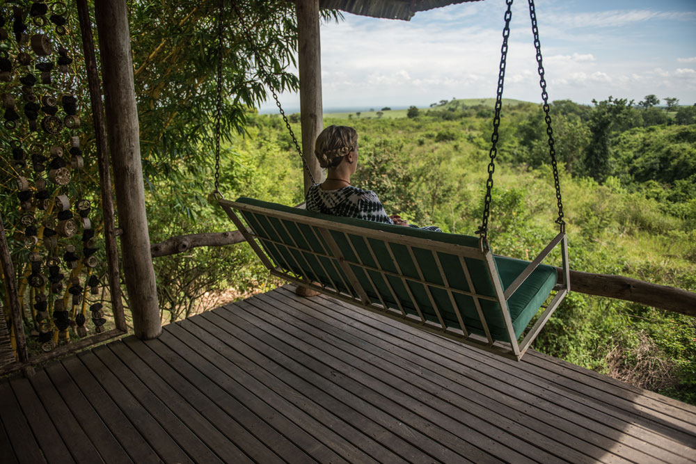 A hanging bench swing on a timber lodge deck, one guest seated on it looking out over dense green bush towards a distant plain