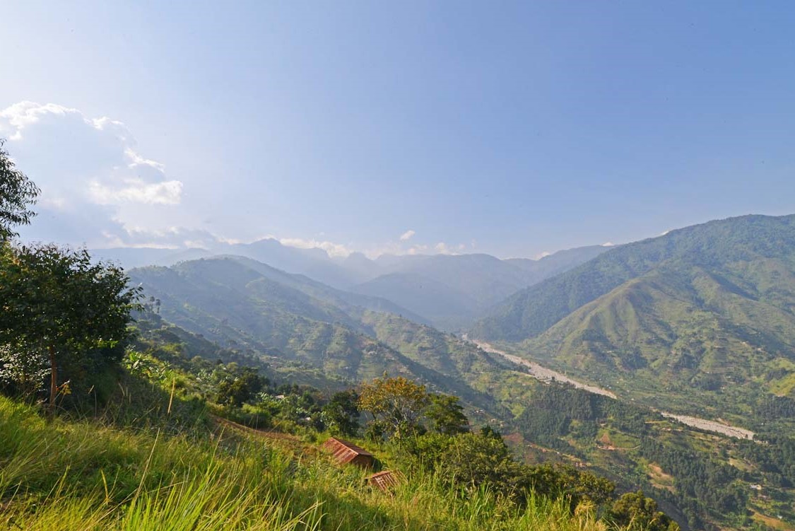 Cultivated green hillsides dropping to a broad valley floor with a pale braided riverbed along it, ranges receding into haze under a high sky