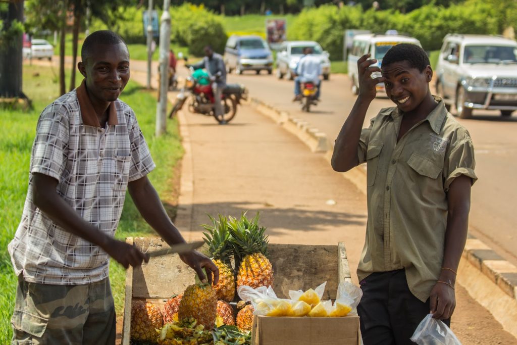 Two street vendors selling pineapples from a roadside stall beside a busy tarmac road