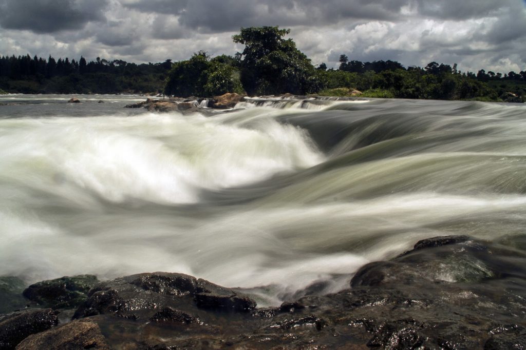 White water pouring over black rock ledges where the Nile leaves Lake Victoria, under a stormy sky